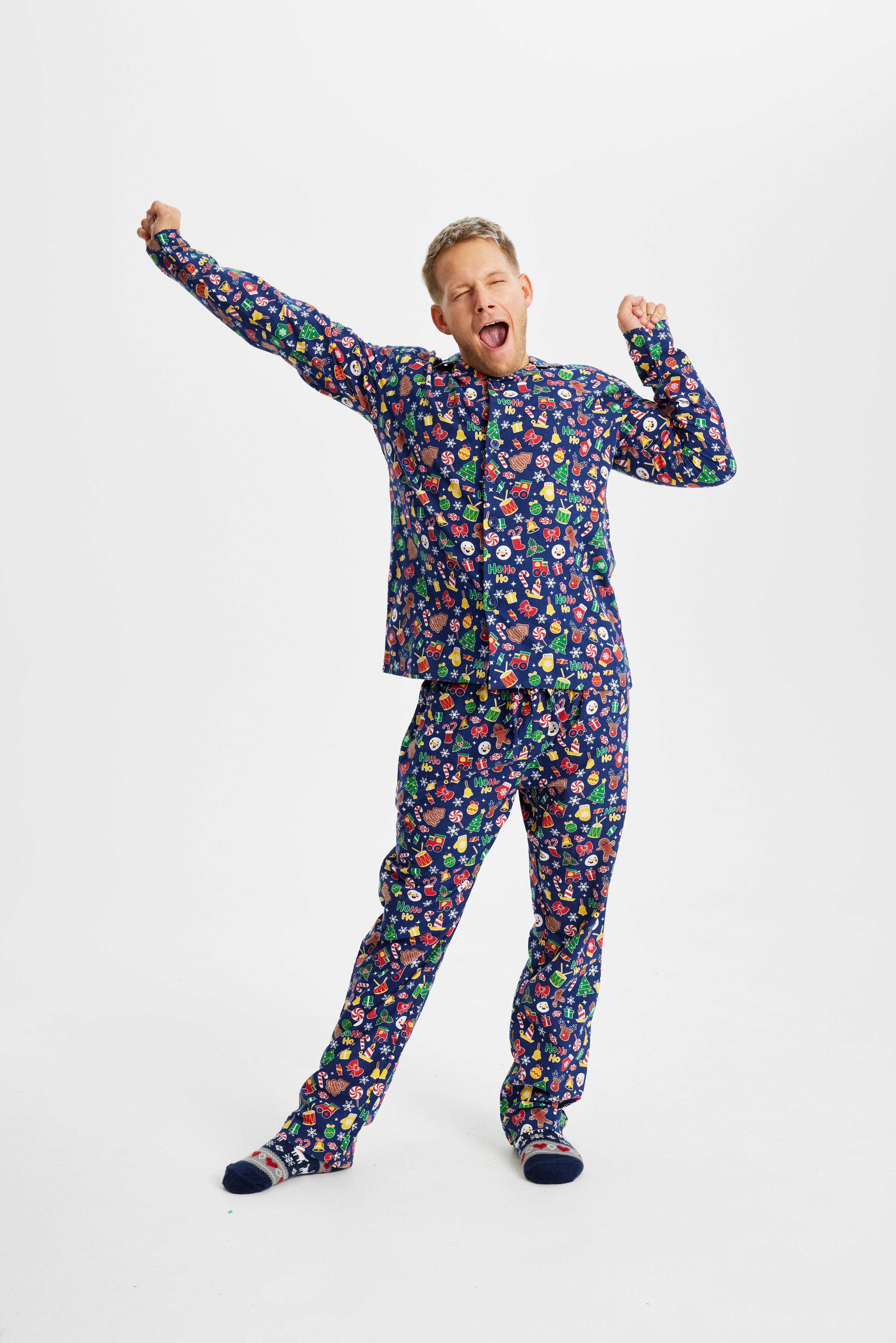 En gabende mand iført en blå julepyjamas med trommer, juleslik, juletræer og julepynt.
